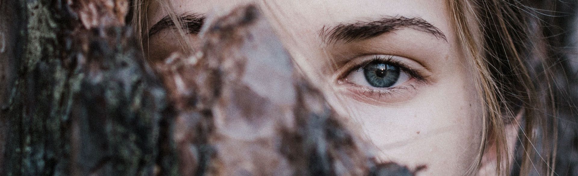 Nahaufnahme eines blauen Auges einer Frau, das hinter einem Baumstamm hervorschaut.